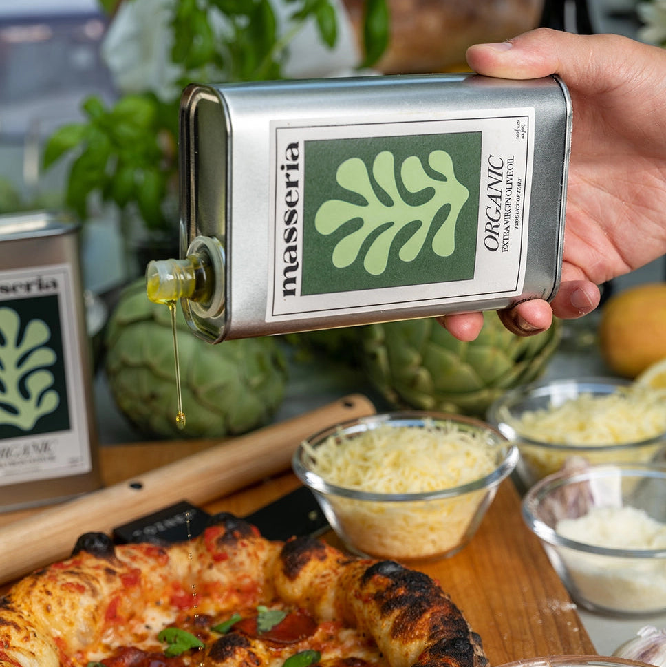 Person holding a Masseria Organic olive oil bottle over a pizza with ingredients on a wooden board.